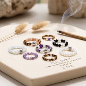 En eksklusiv kollektion af fine, elastiske �delstensringe med facetterede krystaller som ametyst, tiger�je og opalite, smukt pr�senteret p� en lys stenplade med blid r�gelse og t�rrede str� i baggrunden.