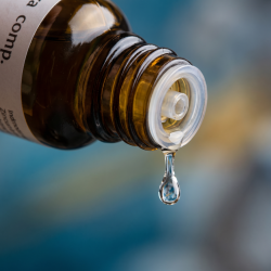 Et krystalklart makrobillede af en enkelt vanddr�be, der falder direkte fra en indbygget dr�bet�ller p� en m�rk ravfarvet glasflaske. Den sl�rede baggrund best�r af dybe, rolige oceanbl� nuancer og glimt af gyldent 'rav'. Lyset er svalt, afstressende og klart.