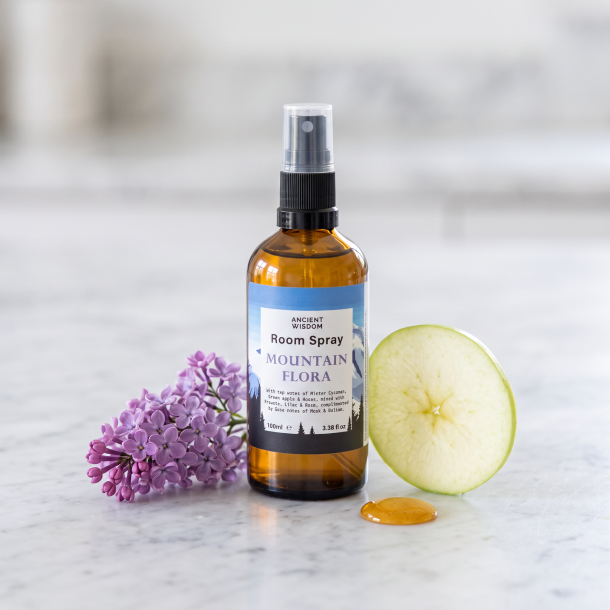N�rbillede af den ravfarvede Ancient Wisdom rumspray glasflaske, st�ende skarpt p� en lys, elegant marmorplade. En frisk gren med lilla syrenblomster, en tynd skive gr�nt �ble og et par dr�ber gylden honning ligger �stetisk placeret omkring flasken i et bl�dt, naturligt lys.