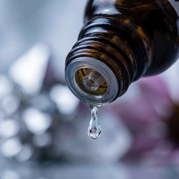 Et krystalklart makrobillede af en vanddr�be, der falder fra den indbyggede dr�bet�ller p� en m�rk ravfarvet glasflaske. Den bl�dt sl�rede baggrund kombinerer elegante, k�lige s�lvgr� og metalliske nuancer med subtile glimt af st�vet lilla, der repr�senterer Solhat (Echinacea) og Argentite krystaller. Lyset er svalt, beroligende og skarpt.