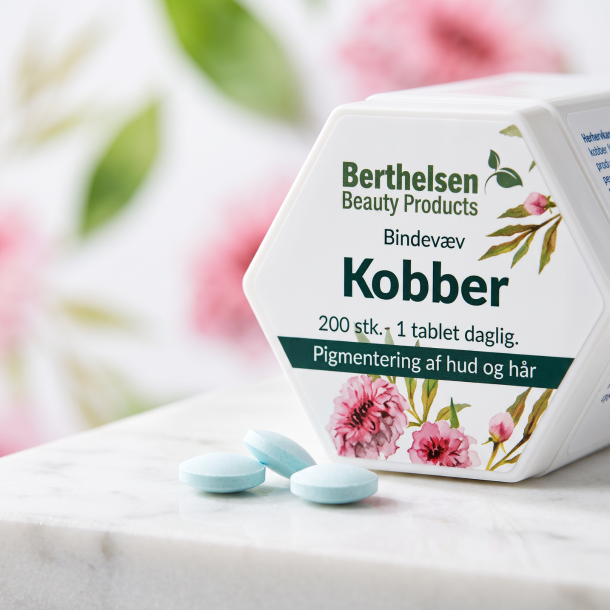 Lysebl� tabletter med kobbergluconat p� en glat overflade ved siden af den sekskantede Berthelsen Kobber d�se med pink blomster.