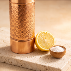 N�rbillede af den hamrede overflade p� Biogans Mosaic kobberflaske. Det gyldne metal reflekterer et bl�dt lys, og foran flasken ligger en halv frisk citron, en lille sk�l med salt og lidt rent vand for at indikere reng�ringsmetoden.