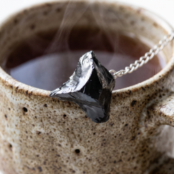 Et n�rbillede af en r�, blank sort obsidian-krystal, der er fastgjort til en s�lvk�de, som hviler mod kanten af et rustikt keramikkrus med varm te.