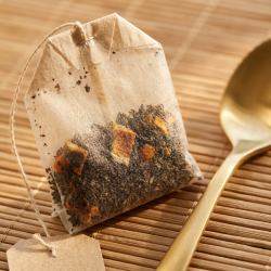 Et lyst n�rbillede af et fint, ubleget tebrev, der ligger ved siden af spidsen af en skinnende, gylden te-ske. Man kan svagt ane de m�rke teblade og orange strejf af citrus igennem det fine papir, der ligger p� en bambusm�tte.