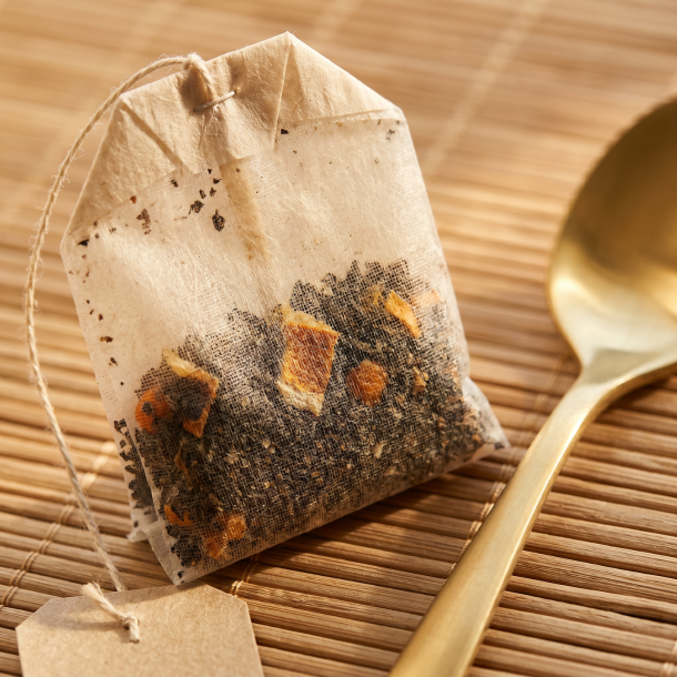 Et lyst n�rbillede af et fint, ubleget tebrev, der ligger ved siden af spidsen af en skinnende, gylden te-ske. Man kan svagt ane de m�rke teblade og orange strejf af citrus igennem det fine papir, der ligger p� en bambusm�tte.