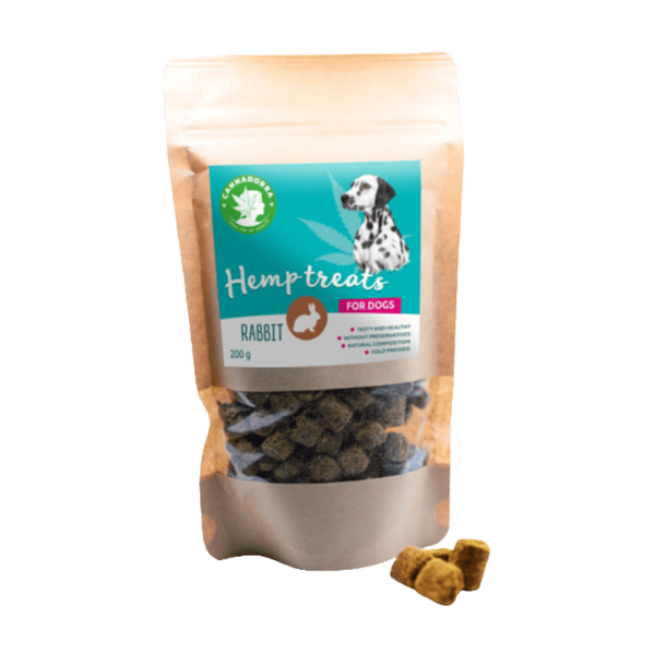 En papirspose med Cannadorra Hemp Treats for Dogs. Etiketten er turkis og viser et billede af en dalmatiner og et lille kaninsymbol. Tre m�rke, koldpressede godbidder ligger foran posen.