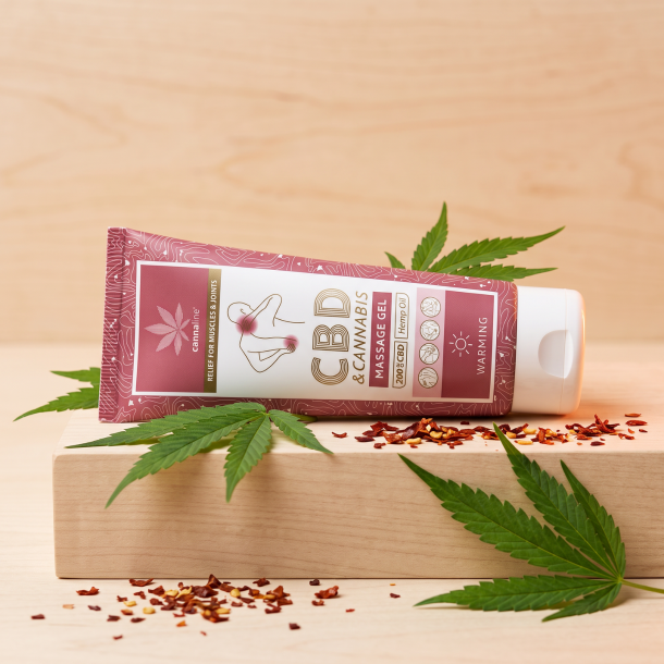 N�rbillede af den st�vede lyser�de/bordeaux Cannaline tube, der st�r skarpt p� en lys overflade af ahorntr�, dekoreret diskret med friske, gr�nne hampeblade og spredte, knuste chiliflager.