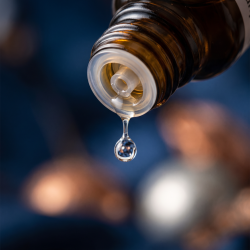 Et krystalklart makrobillede af en vanddr�be, der falder fra den indbyggede dr�bet�ller p� en m�rk ravfarvet glasflaske. Den bl�dt sl�rede baggrund kombinerer dybe, beroligende midnatsbl� nuancer (Cikorie) med varme, metalliske, afsp�ndende glimt af bl�d kobber (Cuprum) og lysende s�lv (Stannum). Lyset er ekstremt klart, varmende, udstr�ler absolut l�sning af indre sp�ndinger og hom�opatisk pr�cision.