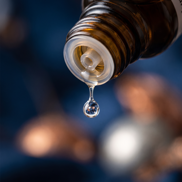 Et krystalklart makrobillede af en vanddr�be, der falder fra den indbyggede dr�bet�ller p� en m�rk ravfarvet glasflaske. Den bl�dt sl�rede baggrund kombinerer dybe, beroligende midnatsbl� nuancer (Cikorie) med varme, metalliske, afsp�ndende glimt af bl�d kobber (Cuprum) og lysende s�lv (Stannum). Lyset er ekstremt klart, varmende, udstr�ler absolut l�sning af indre sp�ndinger og hom�opatisk pr�cision.