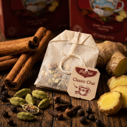 Et sanseligt n�rbillede af et ubleget Yogi tebrev p� m�rkt, rustikt egetr�. Rundt om tebrevet ligger store, tykke kanelst�nger, hele gr�nne kardemommekapsler, t�rrede nelliker, sorte peberkorn og friske stykker ingef�r badet i et varmt lys.