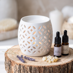 N�rbillede af den hvide, minimalistiske porcel�ns-duftlampe st�ende p� en r� tr�stub i dagslys. Sm� flasker med �teriske olier og en kvist t�rret lavendel ligger ved siden af.