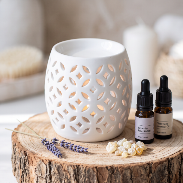 N�rbillede af den hvide, minimalistiske porcel�ns-duftlampe st�ende p� en r� tr�stub i dagslys. Sm� flasker med �teriske olier og en kvist t�rret lavendel ligger ved siden af.