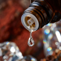 Et krystalklart makrobillede af en vanddr�be, der falder fra den indbyggede dr�bet�ller p� en m�rk ravfarvet glasflaske. Den bl�dt sl�rede baggrund kombinerer varme, dybe rustr�de nuancer med skarpe, krystalklare glimt af lys, der leder tankerne hen p� Vivianit (Jernfosfat) og indre dynamisk varme. Lyset er ekstremt klart, opbyggende og udstr�ler absolut vitalitet, struktur og mineralsk styrke.