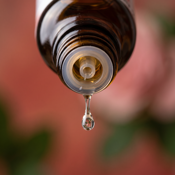 Et krystalklart makrobillede af en vanddr�be, der falder fra den indbyggede dr�bet�ller p� en m�rk ravfarvet glasflaske. Den bl�dt sl�rede baggrund kombinerer varme, omsorgsfulde terrakottar�de og rosa nuancer med blide, dybgr�nne strejf fra Liden N�lde (Urtica urens) og r�de Berberisb�r. Lyset er svalt, feminint, strukturerende og dybt beroligende.