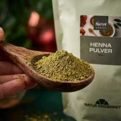 Et krystalklart makrobillede af en rustik, h�ndsk�ret tr�ske, der holder en perfekt bunke af fintmalet, jordagtigt gr�nt plantepulver (henna), klar til at blive blandet. Den bl�dt sl�rede baggrund kombinerer dybe, naturlige gr�nne blade med rige, varme kobber- og mahognir�de farvestrejf. I baggrunden anes silhuetten af den lysegr�nne 1 kg pose fra Natur Drogeriet. Lyset er varmt, indbydende, og udstr�ler absolut botanisk renhed, naturlig sk�nhed og kemifri h�rpleje.