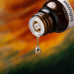 Et krystalklart makrobillede af en vanddr�be, der falder fra den indbyggede dr�bet�ller p� en m�rk ravfarvet glasflaske. Den bl�dt sl�rede baggrund br�nder i varme, gyldne rav- og br�ndte orange nuancer blandet med subtile, dybe skovgr�nne toner, der symboliserer Stormhat (Aconitum) og indre varme. Lyset er klart, opkvikkende og skarpt.