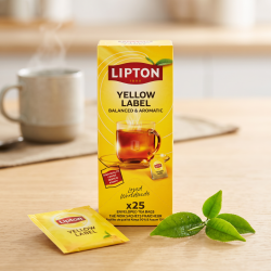 N�rbillede af den klassiske gule Lipton te�ske, der st�r skarpt p� et lyst egetr�sbord. Et enkelt u�bnet, gult tebrev og et par friske, gr�nne teblade ligger dekorativt ved siden af i sollyset.