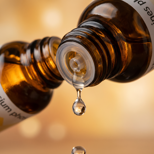 Et krystalklart makrobillede af en vanddr�be, der falder fra den indbyggede dr�bet�ller p� en 1:1 realistisk m�rk ravfarvet glasflaske. Den bl�dt sl�rede baggrund kombinerer varme, beroligende gyldne og bl�de ravfarvede nuancer (Magnesium/Cellesalt 7), der omslutter billedet. Lyset er ekstremt klart, varmende, og udstr�ler absolut indre ro, l�sning af sp�ndinger og hom�opatisk pr�cision.