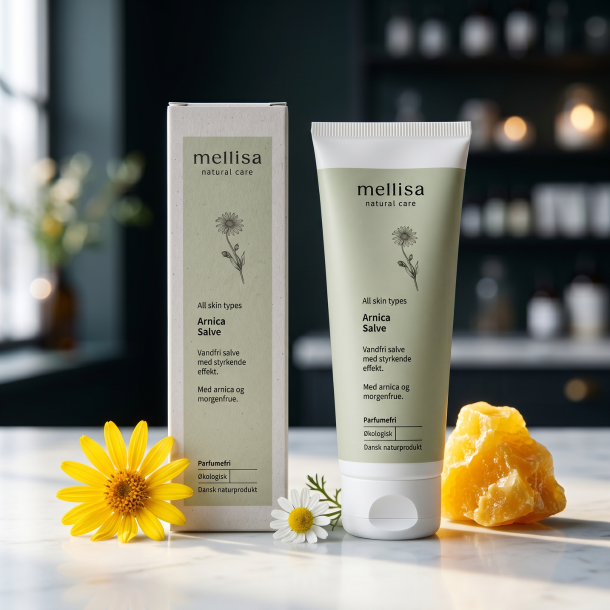 Mellisa Arnica Salve i salviegr�n tube og �ske anrettet p� lyst marmor, omgivet af gul arnica-blomst, kamille og gylden bivoks i et nordisk, eksklusivt milj�.