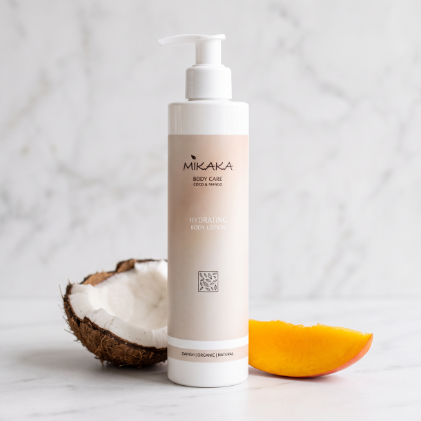 N�rbillede af den hvide Mikaka Coco & Mango flaske, st�ende skarpt p� en lys, mat marmoroverflade. Et stykke br�kket kokosn�d og en lille skive frisk, orange mango ligger utroligt smukt spredt foran flasken i et rent, bl�dt dagslys.