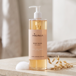 N�rbillede af den gyldne Mikaka Body Wash flaske, st�ende skarpt p� en lys badev�relsesflise af natursten. Et par str� af t�rret, �kologisk havre og en let, cremet s�beboble ligger smukt foran i dagslys.