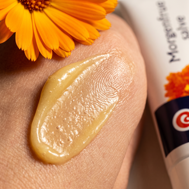 Et l�kkert makrobillede af en rig, cremet og let gylden salve, der sm�res ud p� et glat underlag. I baggrunden ligger knuste, lysende orange kronblade fra en Calendula-blomst. Lyset er varmt og fremh�ver salvens fede, beskyttende og n�rende tekstur.