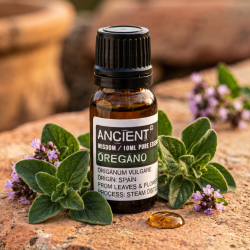 Et n�rbillede af en m�rk glasflaske med �terisk olie. Omkring flasken ligger friske, m�rkegr�nne oreganoblade og sm� lilla blomster badet i varmt middelhavslys p� en rustik flade.