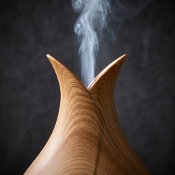 Et n�rbillede af overfladen p� en elegant aroma diffuser i lyst, naturtro egetr�s-look. En fin str�le af kold, hvid damp stiger roligt op fra den buede, tulipanformede top mod en m�rk baggrund.