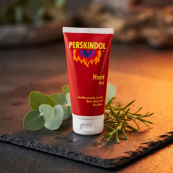 N�rbillede af den knaldr�de Perskindol Heat Gel tube, st�ende skarpt p� en r�, m�rk skiferplade. Et par friske, gr�nne eukalyptusblade og en lille kvist rosmarin ligger omkring tuben i et meget varmt, orange aftenlys, der understreger varmeeffekten.