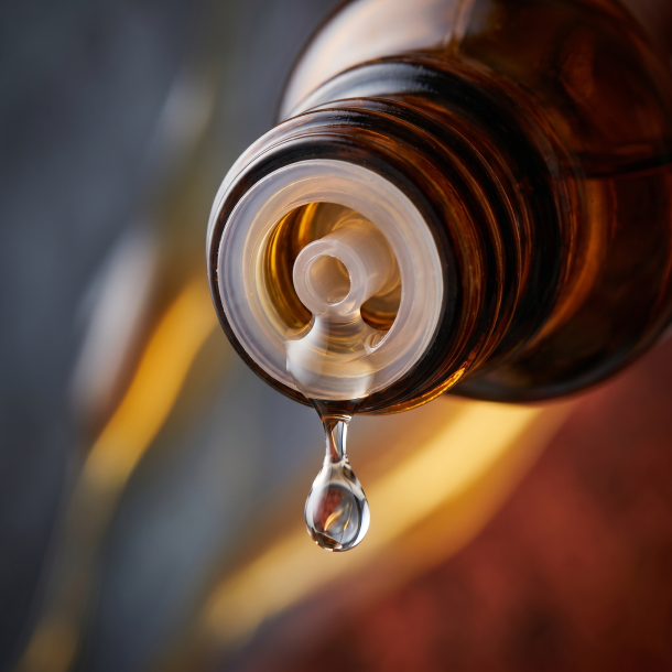 Et krystalklart makrobillede af en vanddr�be, der falder fra den indbyggede dr�bet�ller p� en m�rk ravfarvet glasflaske. Den bl�dt sl�rede baggrund kombinerer dybe, m�rke skifergr� nuancer fra bly (Plumbum) med varme, lysende, gyldne strejf af flydende honning og rustr�d skovmyre (Formica). Lyset er utroligt klart, bl�dg�rende og udstr�ler absolut indre varme, strukturel opl�sning og hom�opatisk pr�cision.