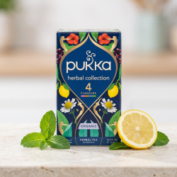 N�rbillede af den dyb bl� og gyldne Pukka Herbal Collection te�ske st�ende skarpt p� en lys stenoverflade. Friske gr�nne mynteblade og en skive citron ligger smukt spredt foran i dagslys.