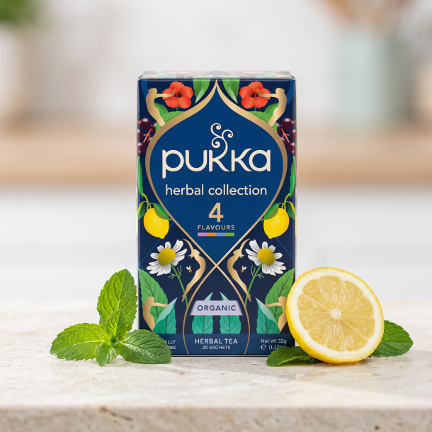 N�rbillede af den dyb bl� og gyldne Pukka Herbal Collection te�ske st�ende skarpt p� en lys stenoverflade. Friske gr�nne mynteblade og en skive citron ligger smukt spredt foran i dagslys.