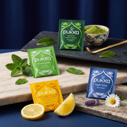 Fire farverige Pukka tebreve pr�senteret p� lyse stenplader. Den gr�nne Supreme Matcha og lysegr�nne Three Mint ligger �verst, mens den gule Lemon & Ginger og bl� Night Time ligger nederst, omgivet af friske citroner, mynte, kamille og matcha-pulver.