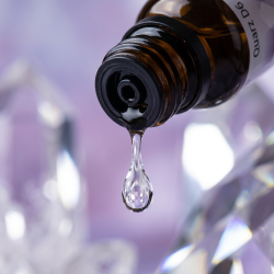 Et krystalklart makrobillede af en vanddr�be, der falder fra den indbyggede dr�bet�ller p� en 1:1 realistisk m�rk ravfarvet glasflaske. Den bl�dt sl�rede baggrund kombinerer sarte, k�lige lavendelnuancer med intense, krystallinske glimt af rent, hvidt lys og geometriske prisme-refleksioner (Quarz/Kisel). Lyset er ekstremt klart, opbyggende, og udstr�ler absolut strukturel formkraft, indre klarhed og hom�opatisk pr�cision.