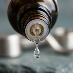 Et krystalklart makrobillede af en vanddr�be, der falder fra den indbyggede dr�bet�ller p� en m�rk ravfarvet glasflaske. I den bl�dt sl�rede baggrund anes subtile, metalliske strejf af s�lv og rolige, jordn�re gr�-bl� nuancer, der symboliserer stabilitet, struktur og grounding. Lyset er klart og fokuseret.