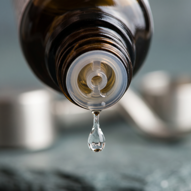 Et krystalklart makrobillede af en vanddr�be, der falder fra den indbyggede dr�bet�ller p� en m�rk ravfarvet glasflaske. I den bl�dt sl�rede baggrund anes subtile, metalliske strejf af s�lv og rolige, jordn�re gr�-bl� nuancer, der symboliserer stabilitet, struktur og grounding. Lyset er klart og fokuseret.