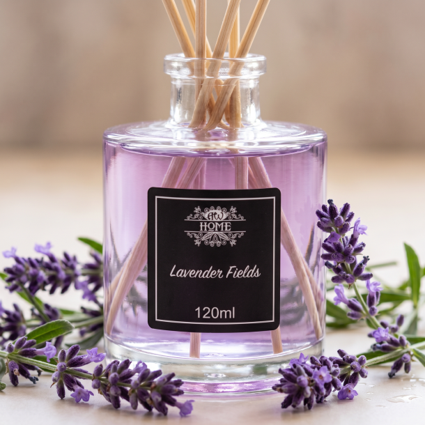 Et n�rbillede af en elegant glasflaske med duftpinde. Sart lilla v�ske i flasken omgivet af friske, vilde lavendelkviste p� en lys overflade med bl�dt lys.