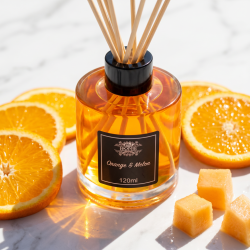 Et n�rbillede af en elegant glasflaske med duftpinde. En lysende, frugtig orange v�ske i flasken omgivet af friske, saftige appelsinskiver og vandmelon p� et lyst marmorbord.