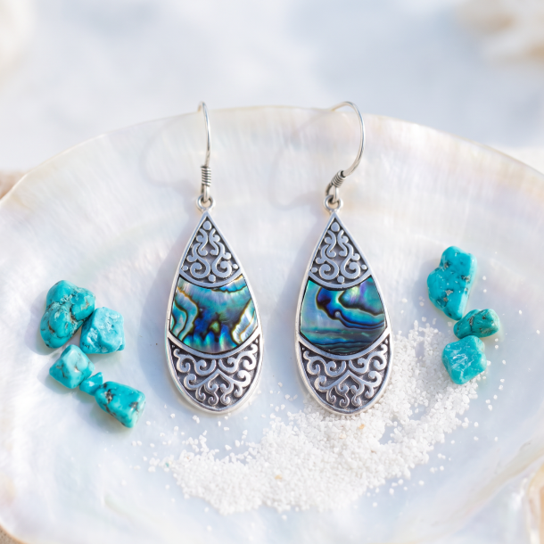 N�rbillede af de dr�beformede Teardrop Oval s�lv�reringe, der hviler smukt p� et stykke lyst og poleret perlemor. Smykkets sirlige s�lvm�nster fanger lyset skarpt. Omkring �reringene ligger en lille, elegant turkis krystal og lidt fint, hvidt strandsand for at understrege havtemaet i et bl�dt dagslys.