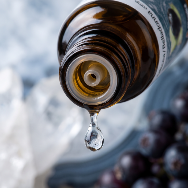 Et krystalklart makrobillede af en vanddr�be, der falder fra den indbyggede dr�bet�ller p� en m�rk ravfarvet glasflaske. Den bl�dt sl�rede baggrund kombinerer elegante, k�lige krystal-hvide og skiferbl� toner (Quartz) med dybe, m�rke b�r-nuancer, der symboliserer Atropa belladonna. Lyset er svalt, struktureret og beroligende.