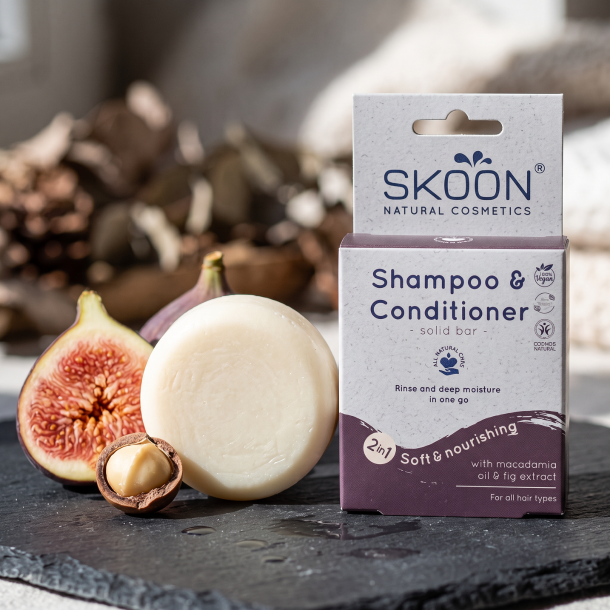N�rbillede af den cremehvide, faste Skoon shampoo bar, der ligger smukt ved siden af sin lilla �ske p� en rustik skiferplade. En halv, frisk figen og en kn�kket macadamian�d ligger �stetisk placeret foran baren i et bl�dt dagslys.