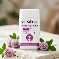 N�rbillede af den hvide og lilla UniKalk d�se, der st�r skarpt p� en lys stenoverflade. Friske lilla r�dkl�verblomster og gr�nne blade ligger smukt spredt omkring den i et bl�dt dagslys.
