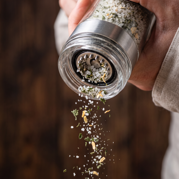 Makrobillede af en saltkv�rn i aktion, der drysser friske, sm�kornede saltkrystaller og t�rrede suppeurter ned i en varm suppe.