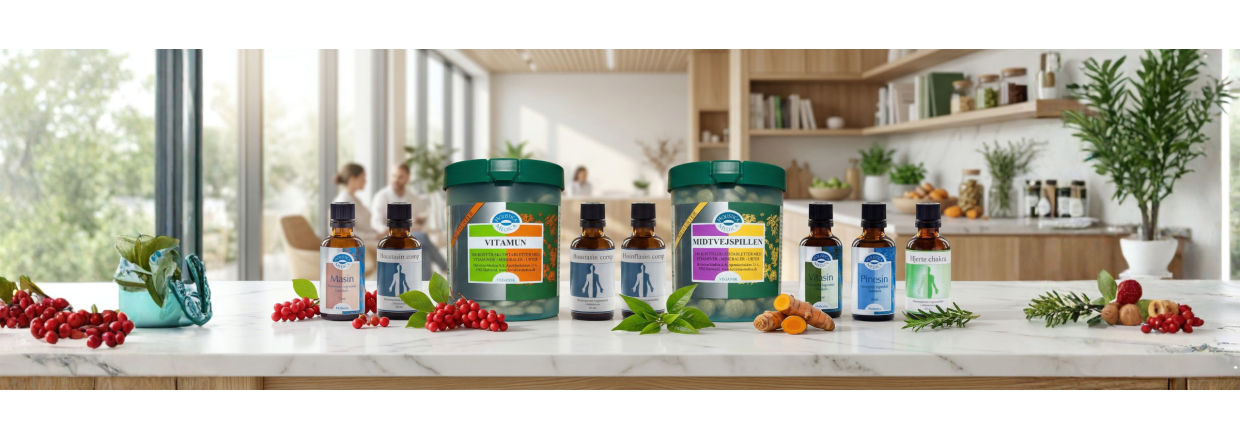 Panorama-banner der viser et bredt sortiment af Holistica-Medica produkter, herunder Midtvejspillen og Vitamun d�ser samt amberfarvede dr�beflasker som Masin, opstillet p� en lys marmorbordplade i et moderne, sollyst k�kkenmilj� med friske urter og b�r.